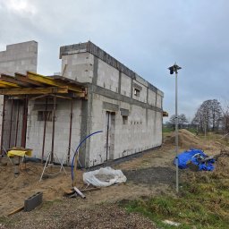 Usługi ogólnobudowlane Groździej Arkadiusz - Budowa domu z betonu komórkowego w trakcie realizacji. Widoczne ściany, szalunki i podpory. Na zewnątrz piasek, narzędzia i materiały budowlane. Pochmurne niebo w tle.