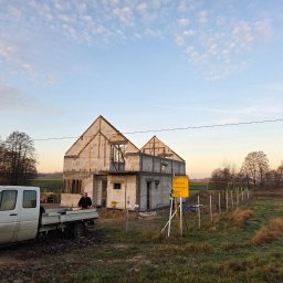 Usługi ogólnobudowlane Groździej Arkadiusz - Wznoszony dom z białego bloczka, widoczny z boku, z częściowo ukończoną konstrukcją dachu. Na pierwszym planie biały samochód dostawczy i pracownik w pomarańczowych rękawicach.