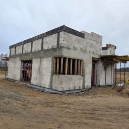 Usługi ogólnobudowlane Groździej Arkadiusz - Surowy stan domu z betonu komórkowego w trakcie budowy. Widoczne szalunki okienne i zbrojenia. Na pierwszym planie ziemia, w tle drzewa i pochmurne niebo.