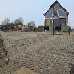 Usługi ogólnobudowlane Groździej Arkadiusz - Ułożona szara ażurowa kostka brukowa na podjeździe do domu w trakcie budowy. Widoczne materiały budowlane i ogrodzenie.