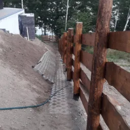 Drewniane ogrodzenie z bali i desek w surowym stylu, z betonową ażurową nawierzchnią i skarpą ziemną w tle, widok z dołu.