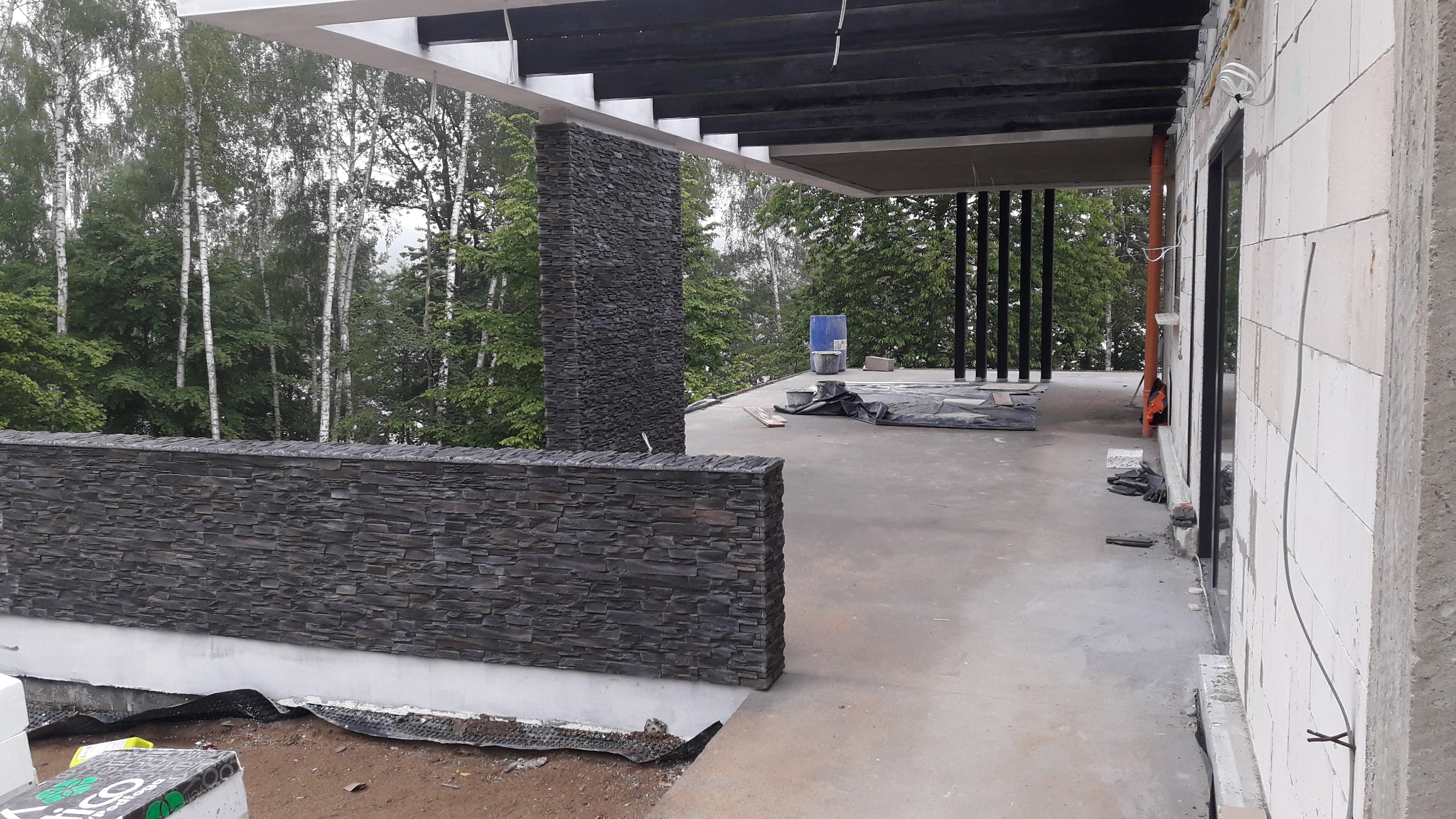 Wznoszona ściana z kamienia elewacyjnego na tarasie domu w stanie surowym otwartym, widoczne materiały budowlane i zieleń w tle.