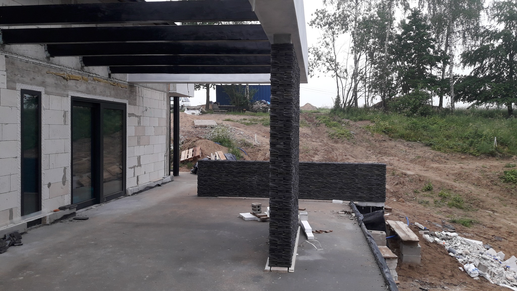 Niedokończony taras z czarną pergolą i kamienną okładziną słupa, widoczne bloczki betonowe i przygotowany teren zielony w tle.