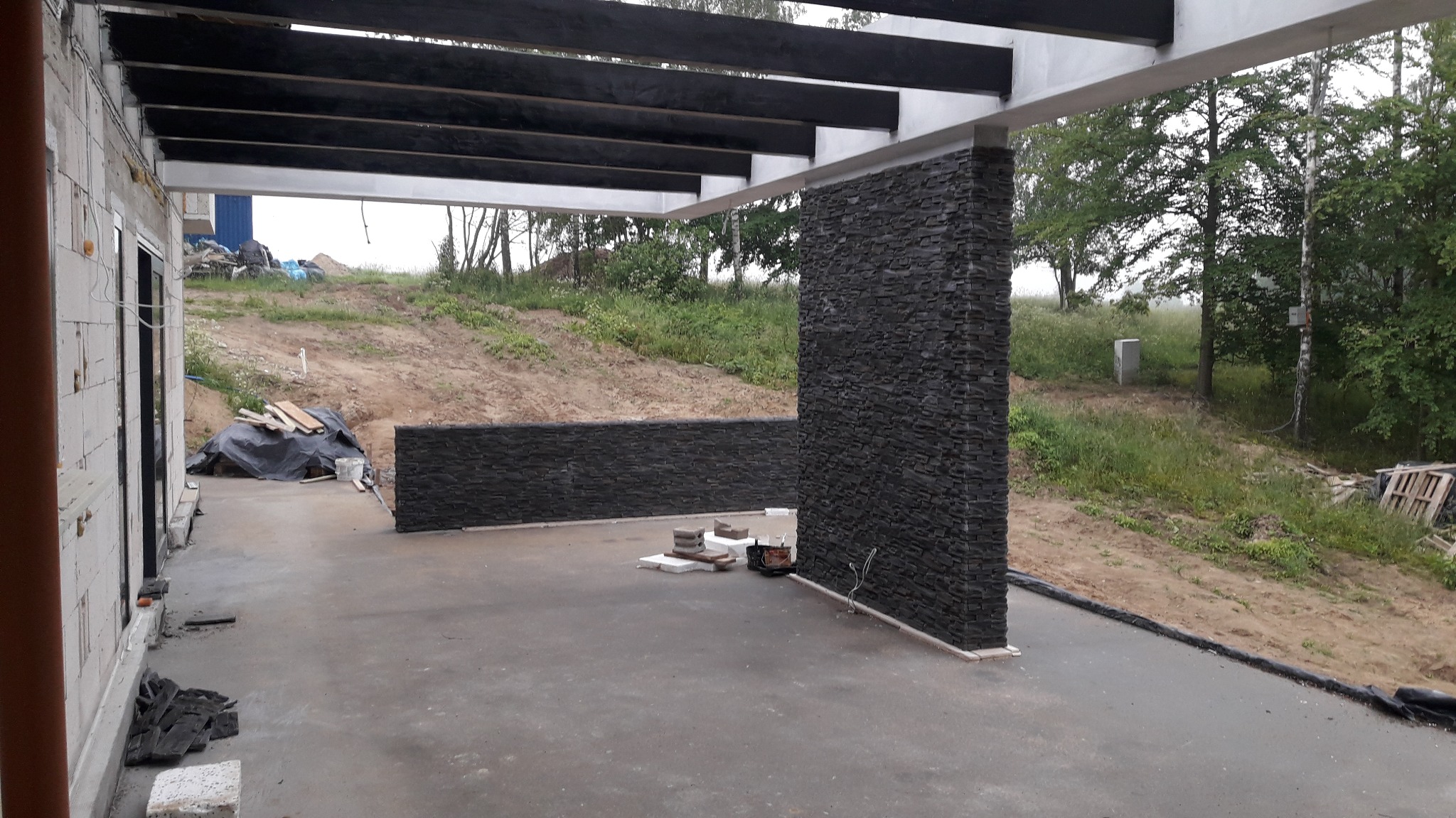 Zadaszony taras w trakcie wykańczania, z fragmentami ścian obłożonych czarnym kamieniem dekoracyjnym, widoczne materiały budowlane i narzędzia na betonowej posadzce, w tle trawiasty teren.