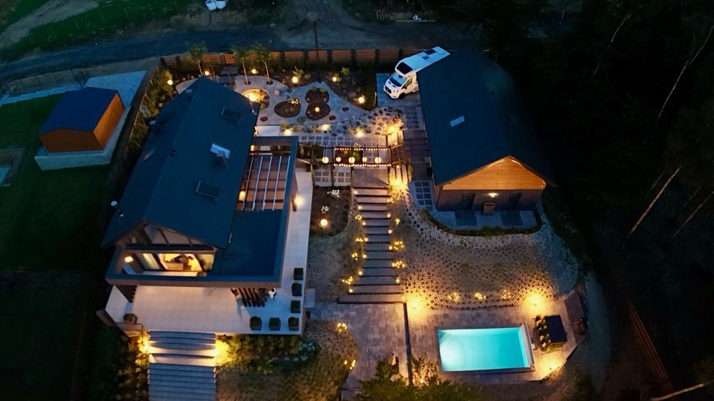 Nocny widok z lotu ptaka na oświetlony dom z basenem, ogrodem i kamperem. Nowoczesna architektura, kostka brukowa i dekoracyjne oświetlenie tworzą luksusowy krajobraz.