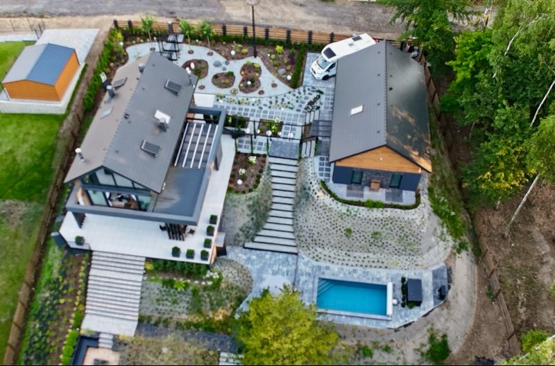 Widok z lotu ptaka na nowoczesny dom z basenem, tarasem i starannie zaprojektowanym ogrodem z kamiennymi ścieżkami i roślinnością. Na posesji zaparkowany kamper.