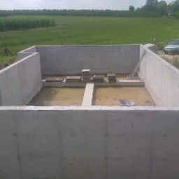 Wylany fundament domu z widocznymi deskami wewnątrz, na tle zielonego krajobrazu i częściowo widocznym samochodem osobowym.