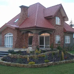 Elegancki dom z cegły klinkierowej z czerwonym dachem, widok z ogrodu. Uwagę przyciąga kamienny murek okalający rabatę z roślinami.