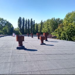 BZIUK GARAGE MARCIN ŁUKASIEWICZ - Płaski dach pokryty papą z widocznymi kominami wentylacyjnymi. W tle zieleń drzew i błękitne niebo. Widoczne łączenia papy. Kominki w kolorze rdzawym.