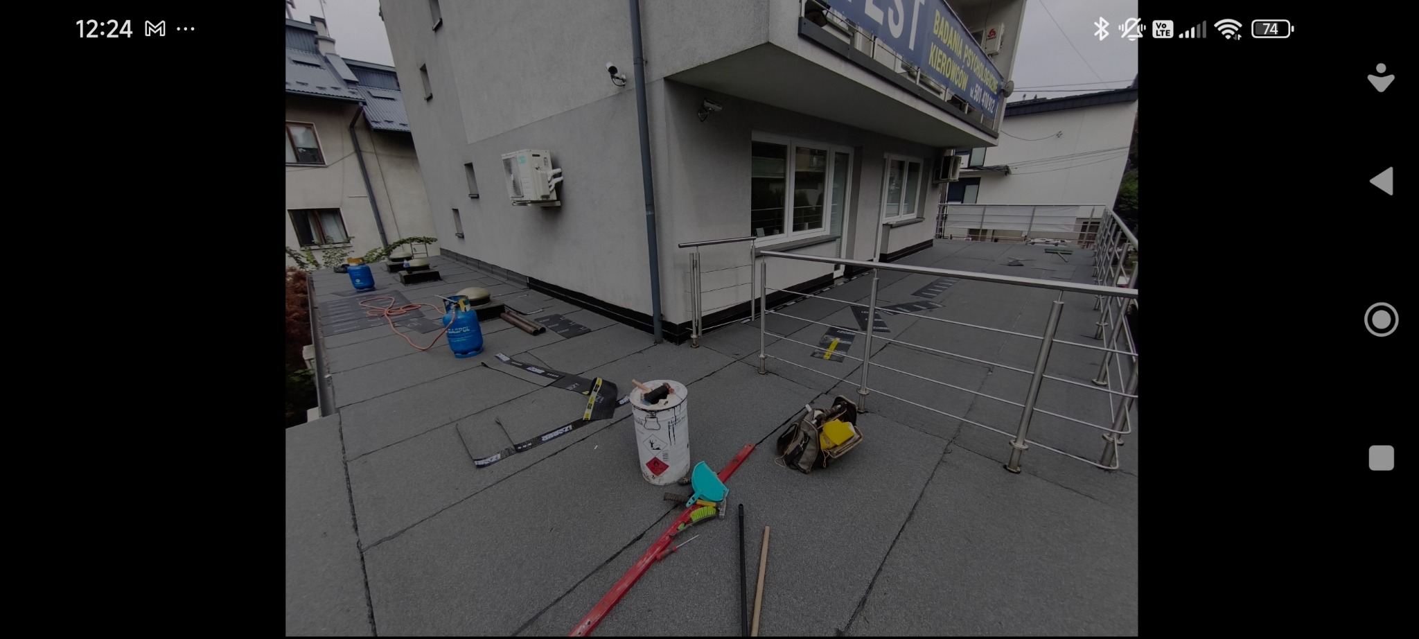 Płaski dach budynku z narzędziami i butlami gazowymi, widok z góry. Na dachu widoczne materiały izolacyjne i metalowe barierki. Prace dekarskie w toku.