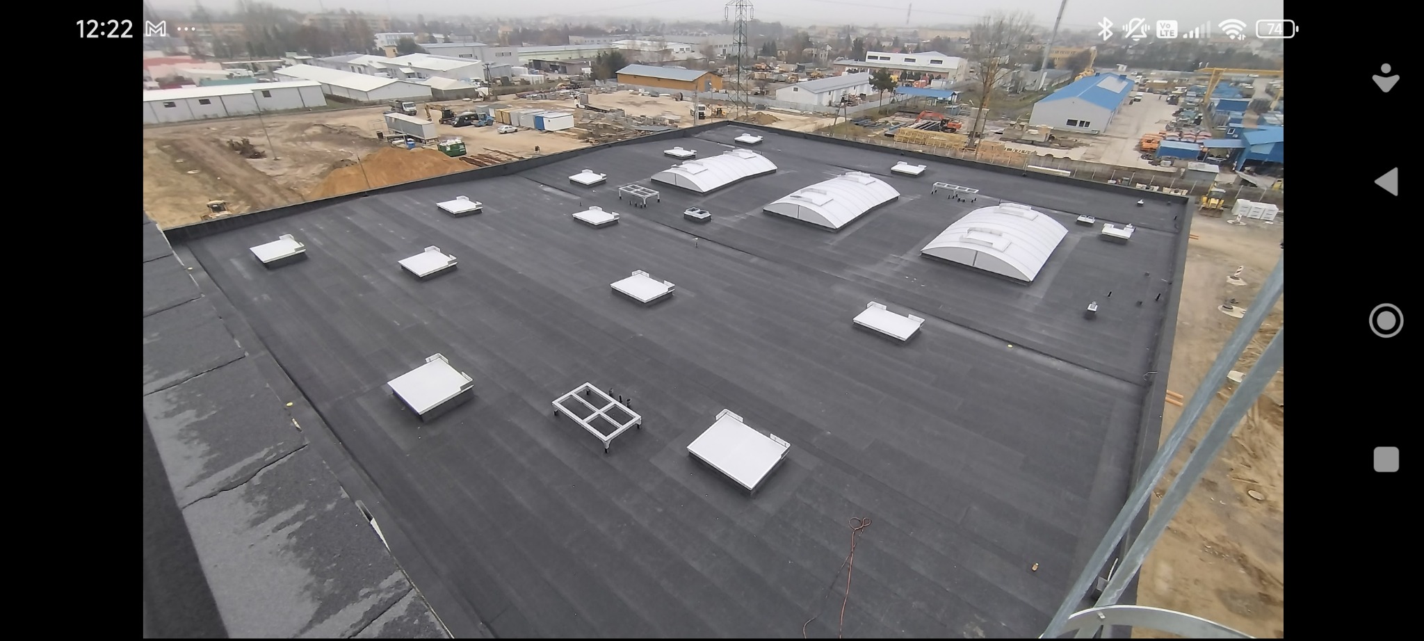 Czarny, płaski dach przemysłowy z świetlikami i elementami konstrukcyjnymi. Widok z góry, z elementami rusztowania w prawym dolnym rogu. Tło stanowią budynki przemysłowe.