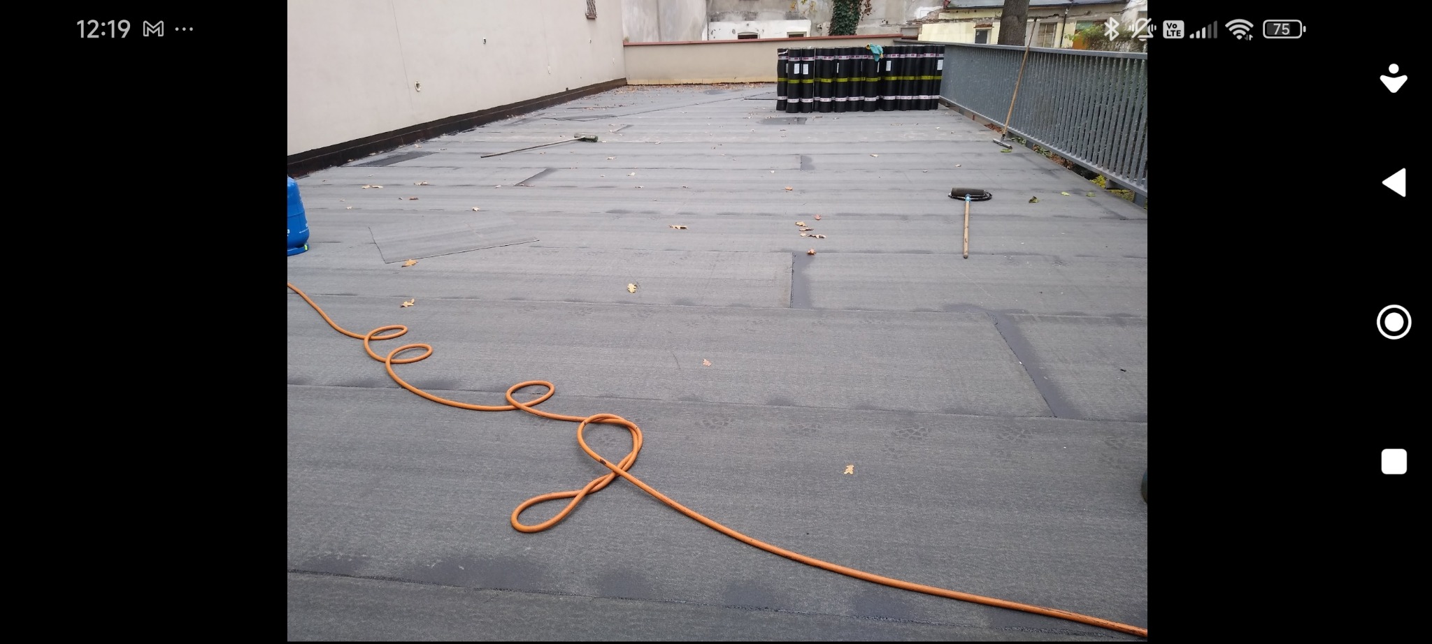 Wykończony dach pokryty papą termozgrzewalną. Widoczne zgrzewy, narzędzia dekarskie i rolki papy w tle. Pomarańczowy wąż na pierwszym planie.