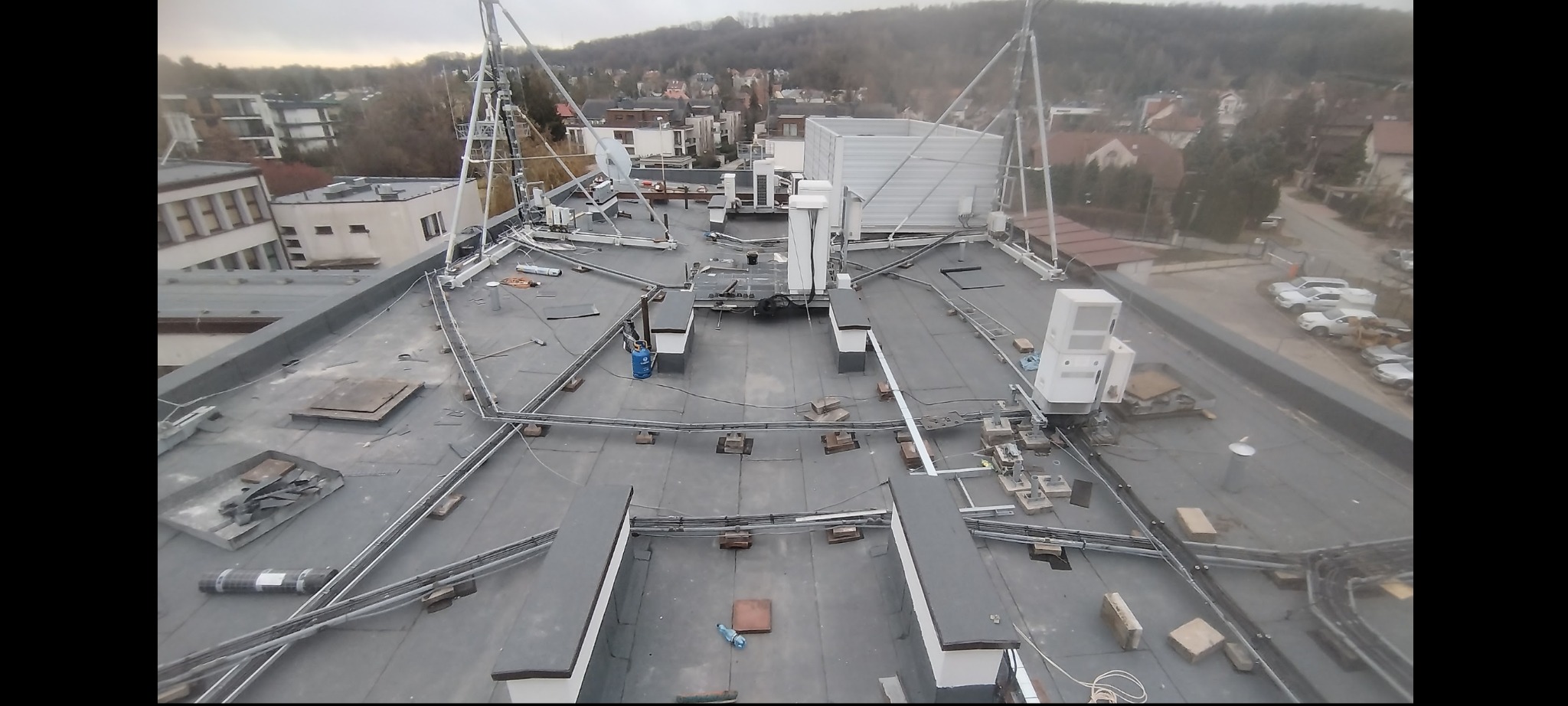 Widok z góry na płaski dach budynku z instalacjami, przewodami i elementami konstrukcyjnymi. W tle zabudowa miejska i zieleń. Ujęcie z góry, w dzień.