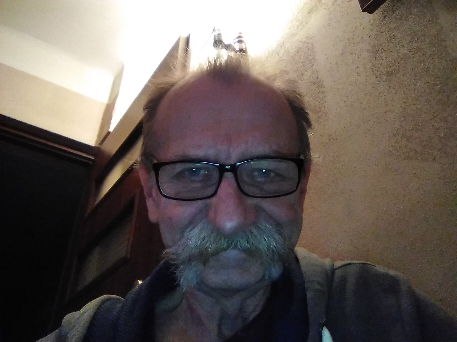 Portret mężczyzny w okularach z wąsami na tle ściany i fragmentu drzwi. Oświetlenie od góry.