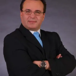 Portret mężczyzny w okularach, czarnym garniturze i błękitnym krawacie, z zegarkiem na lewym nadgarstku, skrzyżowane ręce na tle szarego tła.