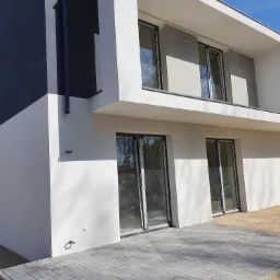 Nowoczesny dom jednorodzinny z szarą elewacją i dużymi oknami, widoczny fragment utwardzonego terenu z kostki brukowej.