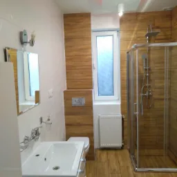 Nowoczesna łazienka po remoncie w Łoniowie z kabiną prysznicową, umywalką z szafką, lustrem i płytkami imitującymi drewno na ścianach i podłodze.