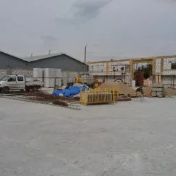 Budowa budynku z białych bloczków: pracownicy na rusztowaniu, zaplecze budowy z materiałami i pojazdami, widok na halę w tle.