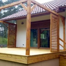 Drewniana pergola z ażurowymi ściankami i schodami przy domu jednorodzinnym, widok z zewnątrz na taras z desek.