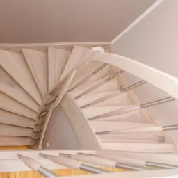 Jasne, drewniane schody spiralne z metalowymi balustradami, widok z góry, oświetlenie punktowe na ścianie.