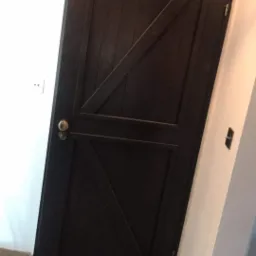 Ciemne, drewniane drzwi wewnętrzne z ukośnymi deskami w stylu rustykalnym, z zamontowaną złotą klamką i widocznymi elementami wykończenia wnętrza.