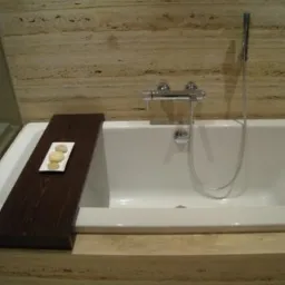 Biała wanna z chromowaną baterią i prysznicem, drewniana półka z trzema kamieniami na boku, ściana wyłożona płytkami imitującymi trawertyn.