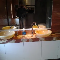 Dwie owalne, marmurkowe umywalki w odcieniach żółci, z chromowanymi bateriami, osadzone na brązowym blacie z marmuru i białych szafkach. W lustrze odbija się osoba robiąca zdjęcie telefonem.