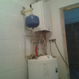 Biały bojler elektryczny z niebieskim zbiornikiem wyrównawczym, zamontowany na ścianie wyłożonej białymi płytkami w pomieszczeniu z widoczną ścianą z cegły w tle.