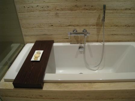 Biała wanna z chromowaną baterią i prysznicem, drewniana półka z trzema kamieniami na boku, ściana wyłożona płytkami imitującymi trawertyn.