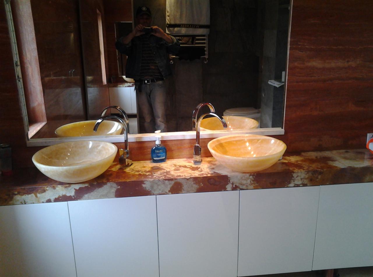 Dwie owalne, marmurkowe umywalki w odcieniach żółci, z chromowanymi bateriami, osadzone na brązowym blacie z marmuru i białych szafkach. W lustrze odbija się osoba robiąca zdjęcie telefonem.