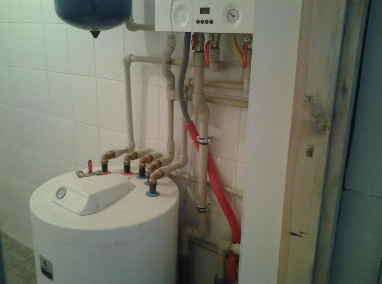 Biały bojler z licznymi rurami i zaworami w pomieszczeniu wyłożonym białymi płytkami, widoczny fragment pieca gazowego na ścianie.