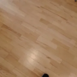 Jasna, drewniana podłoga parkietowa, ułożona w regularny wzór. W rogu widoczny fragment buta. Delikatne oświetlenie podkreśla naturalną strukturę drewna.