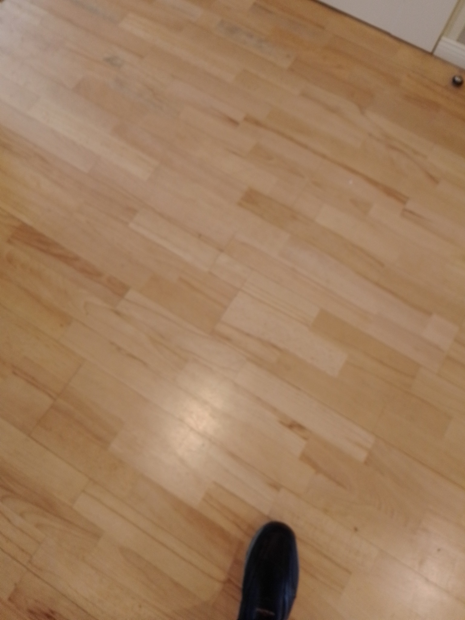 Jasna, drewniana podłoga parkietowa, ułożona w regularny wzór. W rogu widoczny fragment buta. Delikatne oświetlenie podkreśla naturalną strukturę drewna.