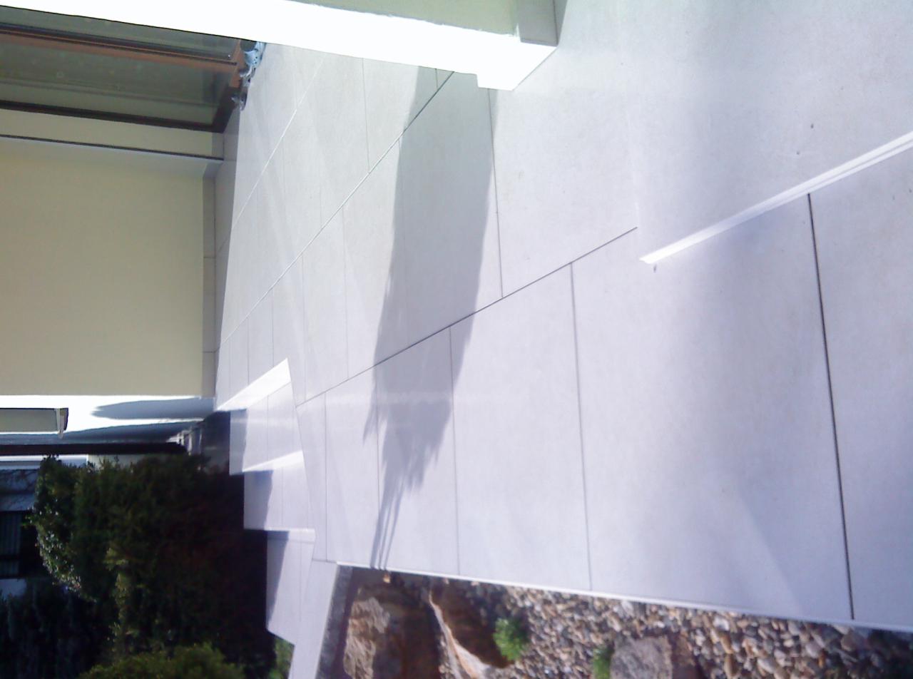 Ułożone jasnoszare płytki na tarasie z widocznym cieniem drzewa, obok rabata z kamykami i zielenią.