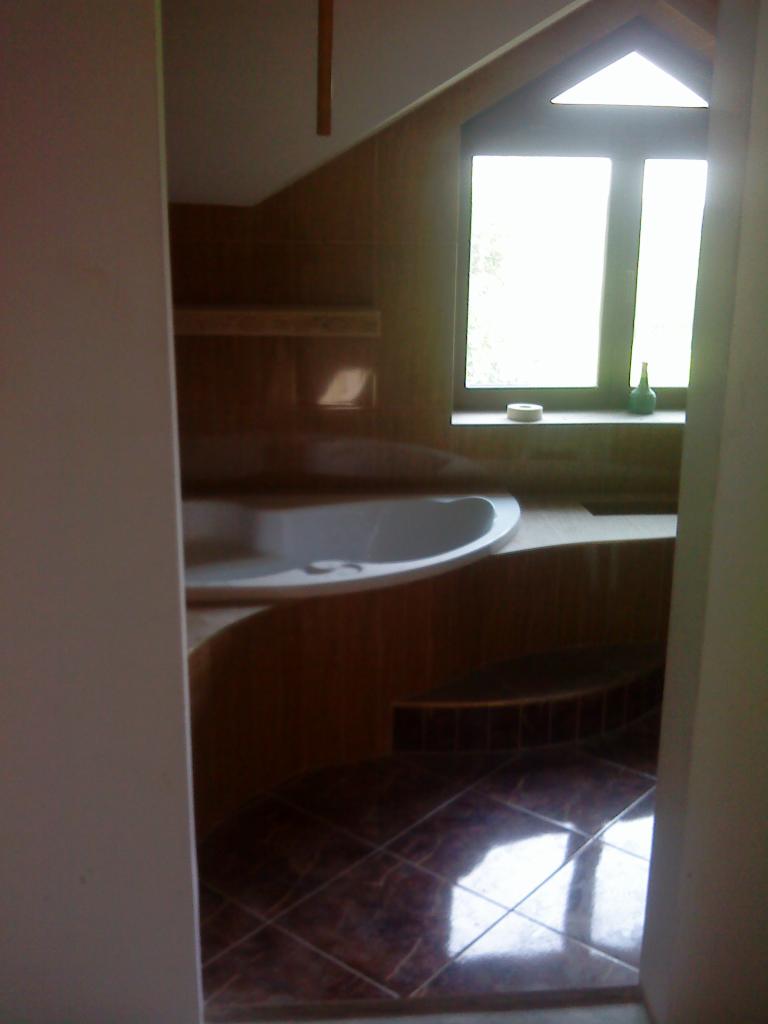 Kompleksowo wykończona łazienka na poddaszu z brązowymi płytkami podłogowymi i wanną w zabudowie, widok zza futryny drzwi.