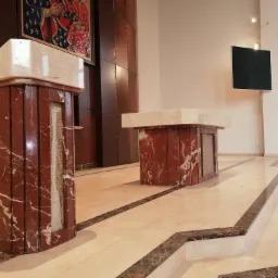 Dwa marmurowe postumenty z blatami z jasnego kamienia w kościele, z mozaiką religijną na ścianie w tle i marmurową posadzką.