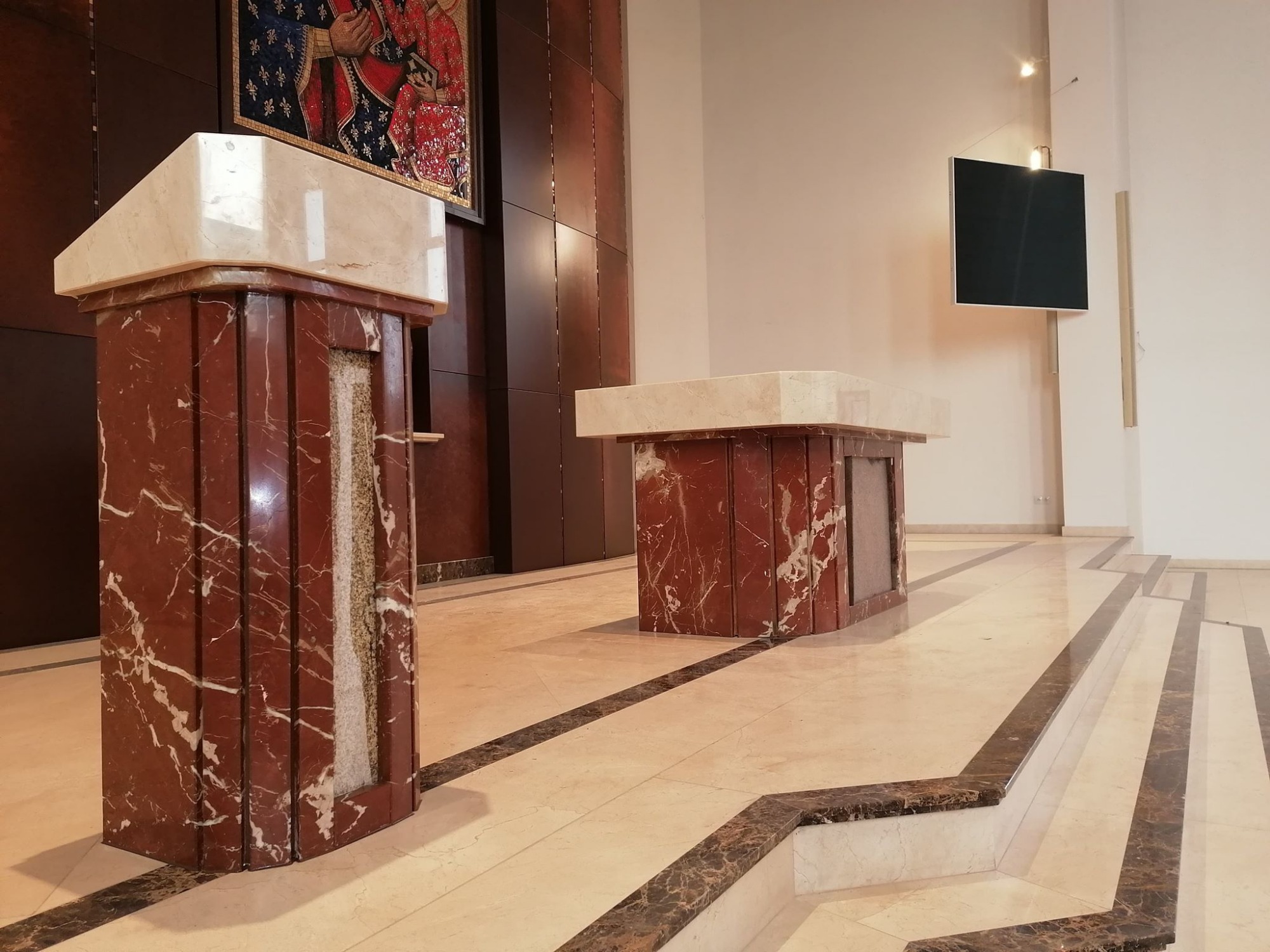 Dwa marmurowe postumenty z blatami z jasnego kamienia w kościele, z mozaiką religijną na ścianie w tle i marmurową posadzką.