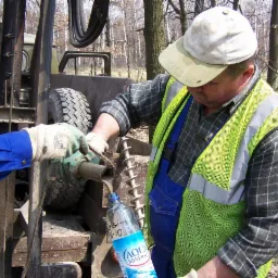 Pracownik w kamizelce BHP pobiera próbkę gruntu do butelki podczas wiercenia. W tle drzewa i maszyna wiertnicza. Wiertło i gleba w kadrze.