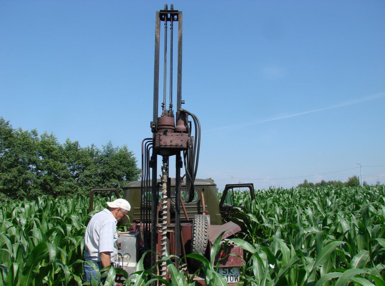 Praca w polu kukurydzy: wiertnica zamontowana na pojeździe, obsługiwana przez mężczyznę w czapce, na tle błękitnego nieba i zieleni.