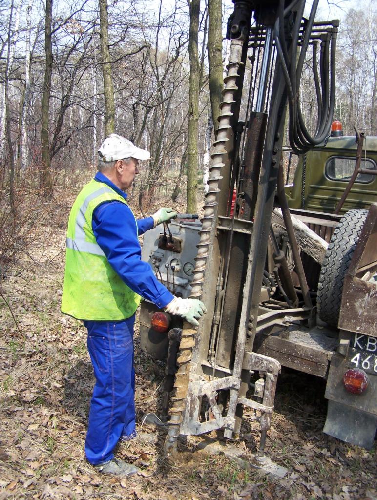 Pracownik w kamizelce obsługuje wiertnicę na tle lasu. Wiertło zagłębia się w ziemię, obok widoczny fragment ciężarówki z numerem rejestracyjnym.