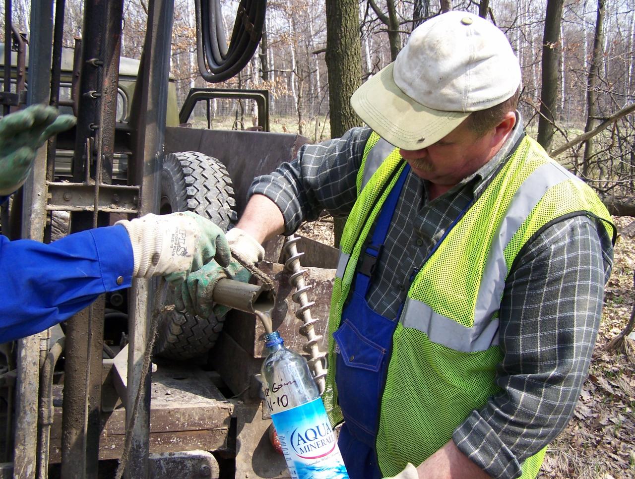 Pracownik w kamizelce BHP pobiera próbkę gruntu do butelki podczas wiercenia. W tle drzewa i maszyna wiertnicza. Wiertło i gleba w kadrze.