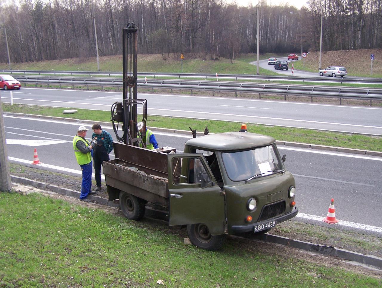 Ekipa przy UAZ-ie z wiertnicą na poboczu drogi, przygotowująca się do odwiertów geologicznych. W tle ruch samochodowy i drzewa.