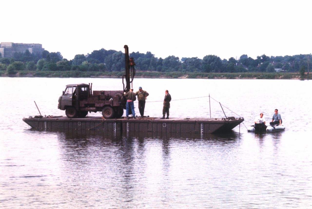 Prace geologiczne na wodzie: platforma z ciężarówką i wiertnicą, grupa osób na platformie i ponton na rzece, budynek w tle. Nietypowe ujęcie.