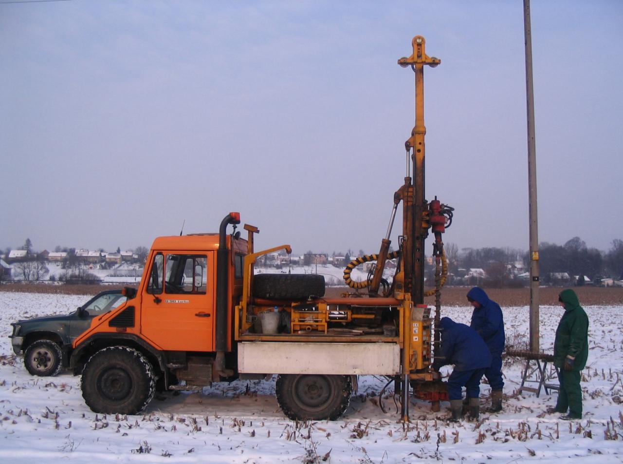 Pomarańczowy samochód ciężarowy z wiertnicą na śniegu, pracownicy w kombinezonach podczas wiercenia, w tle zabudowania.