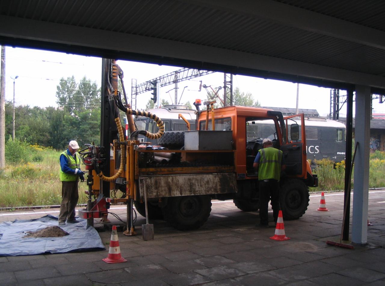 Prace geologiczne przy torach kolejowych: pomarańczowy pojazd specjalistyczny z wiertnicą, dwóch pracowników w kamizelkach odblaskowych, próbka gruntu na folii.