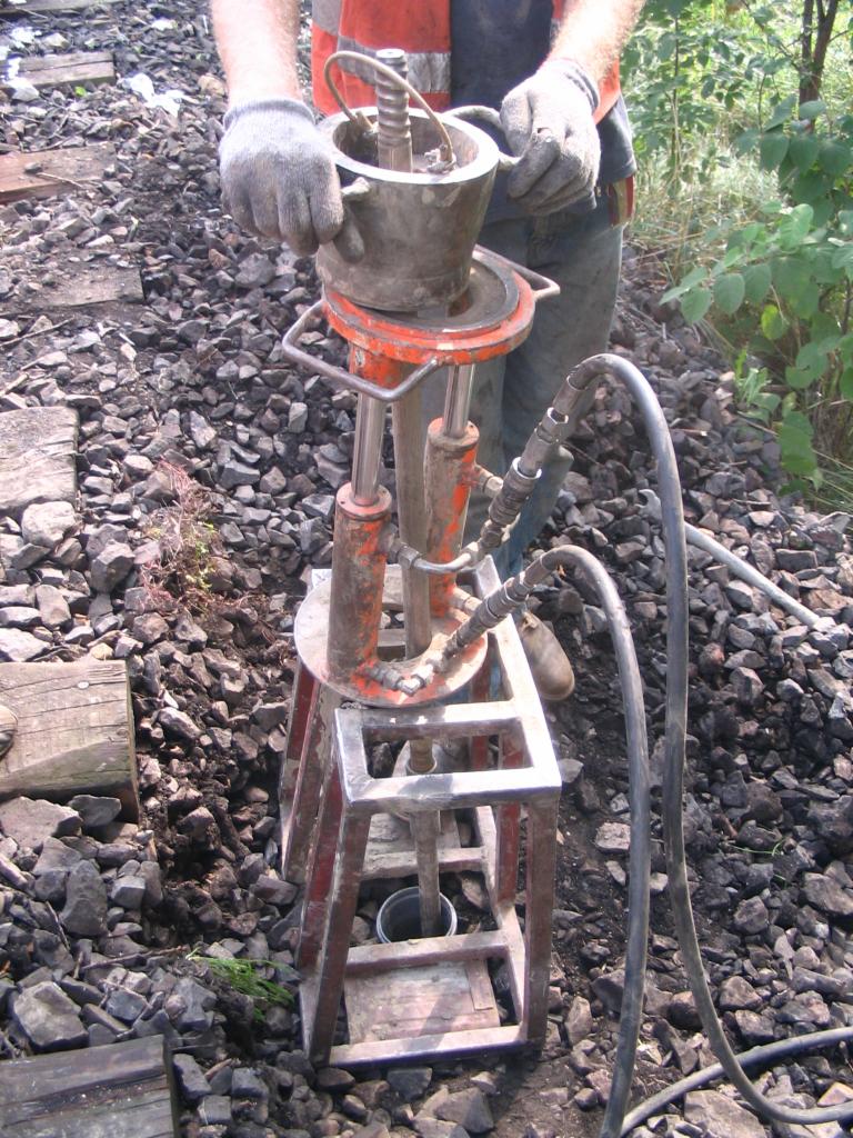 Praca geologiczna: operator w rękawicach obsługuje urządzenie do poboru próbek gruntu na terenie z gruzem i roślinnością.