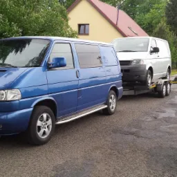 Niebieski Volkswagen Transporter T4 z chromowanym orurowaniem holuje na lawecie białego Transportera T4 na tle wiejskiej drogi i domu.