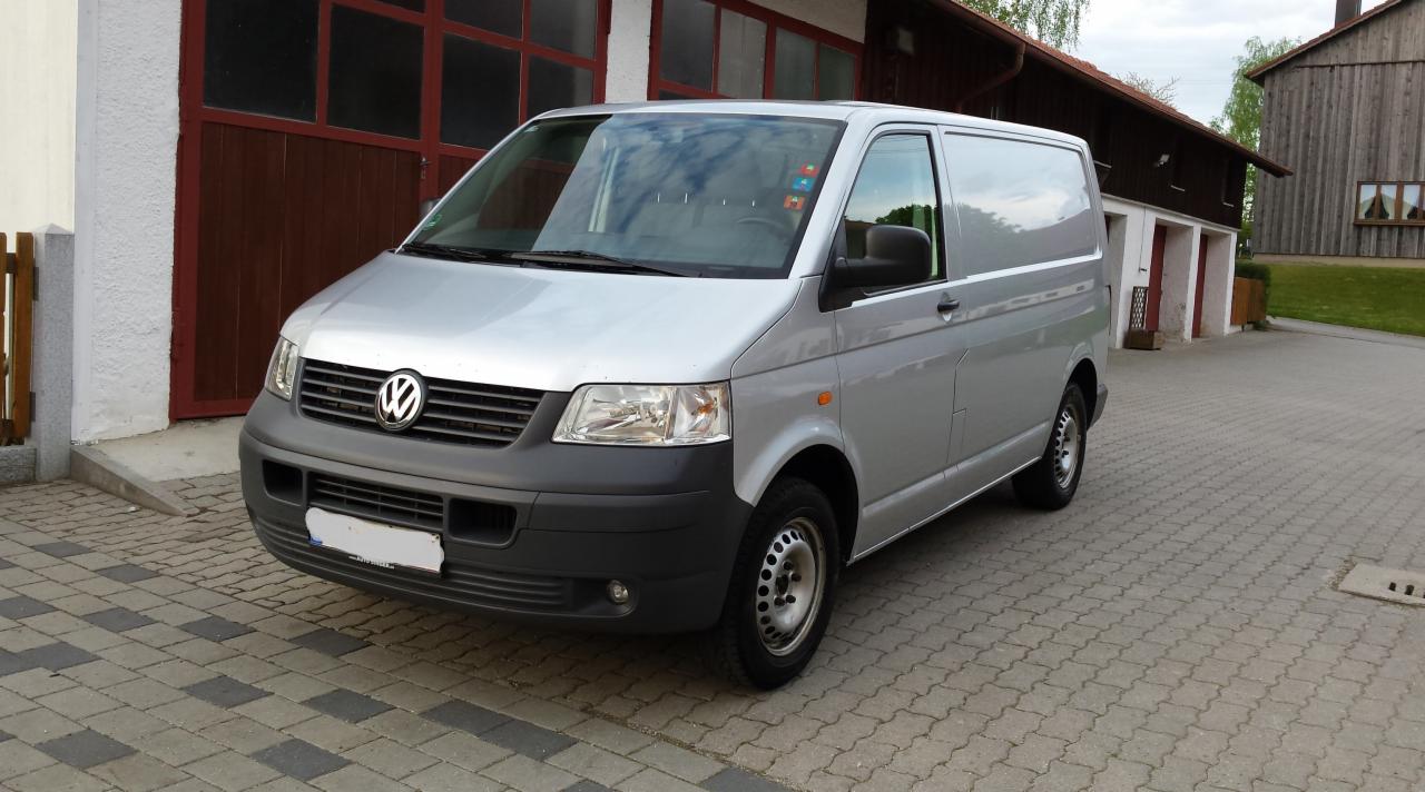 Srebrny Volkswagen Transporter T5, zaparkowany na brukowanej nawierzchni przed budynkiem z czerwonymi drzwiami garażowymi, w pochmurny dzień.
