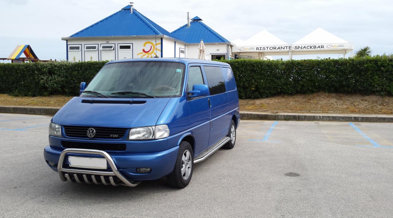 Niebieski Volkswagen Transporter T4 na parkingu przed budynkiem z niebieskim dachem i napisem Restaurante Snackbar. Auto z orurowaniem ochronnym.