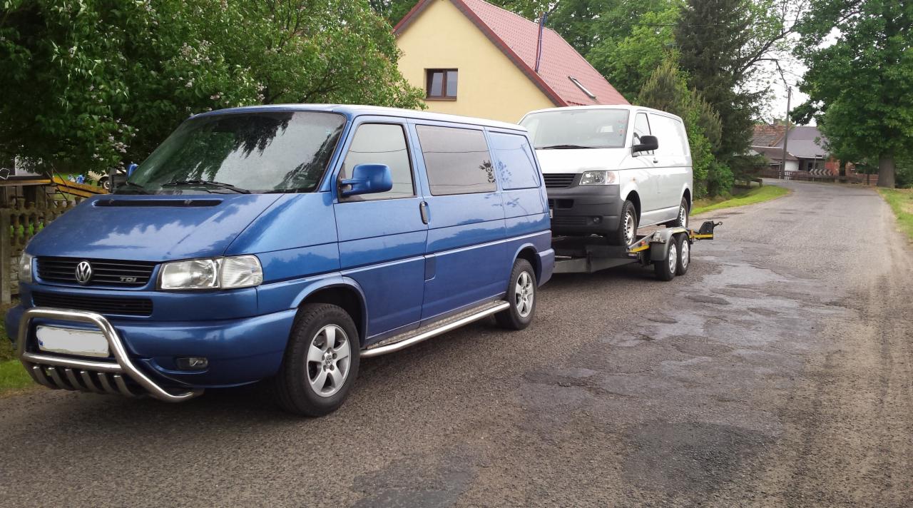 Niebieski Volkswagen Transporter T4 z chromowanym orurowaniem holuje na lawecie białego Transportera T4 na tle wiejskiej drogi i domu.